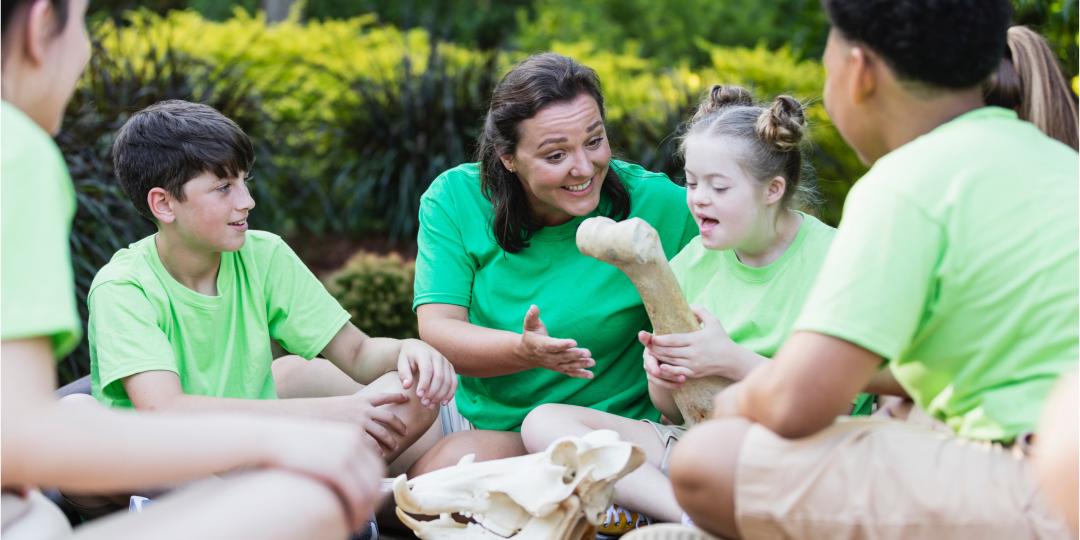 Kind mit einer sichtbaren kognitiven Beeinträchtigung (Trisomie 21)  in einer Gruppe von Kindern und einer erwachsenen Frau im Freien