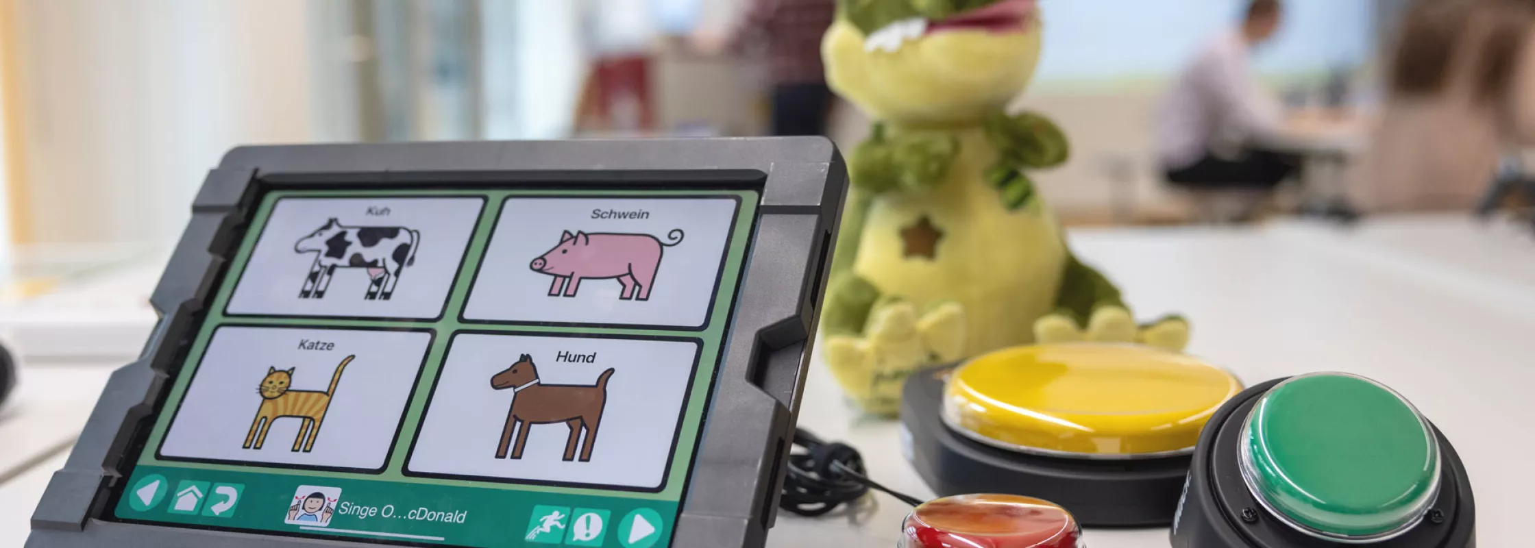 Auf einem Tisch stehen drei unterschiedliche Technologien zur unterstützen Kommunikation. Links ein iPad in einer robusten Halterung mit einem Programm mit Symbolen zur unterstützen Kommunikation. Im Hintergrund sitzt ein Krokodil-Plüschtier welches singen und tanzen kann. Das Krokodil ist an Taster rechts im Bild angeschlossen über welche das Krokodil an und ausgeschaltet werden kann. Die Taster sind zwischen 5 und 10cm gross und sehr robust.