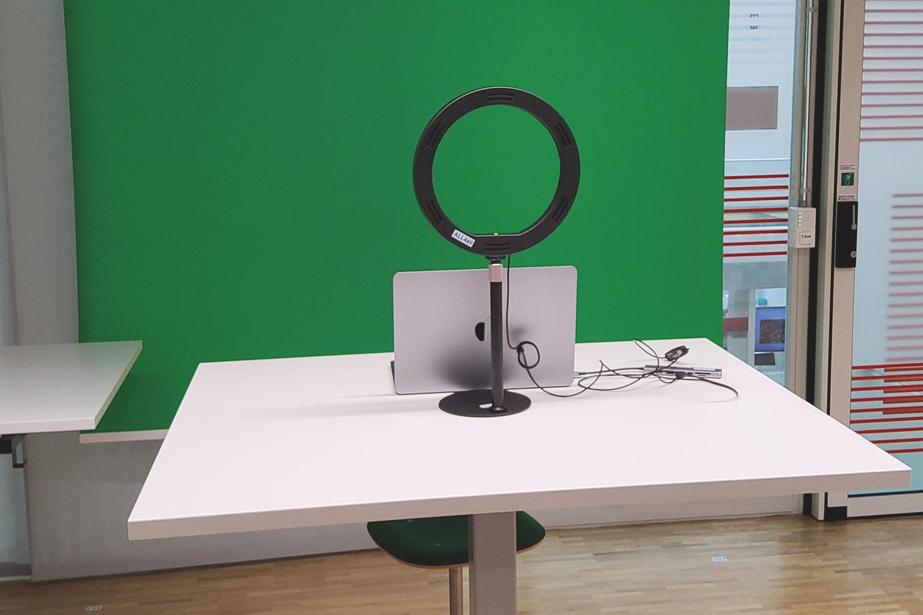 Foto eines Greenscreens mit davor aufgestelltem Tisch auf dem ein Laptop und eine Ringleuchte steht.