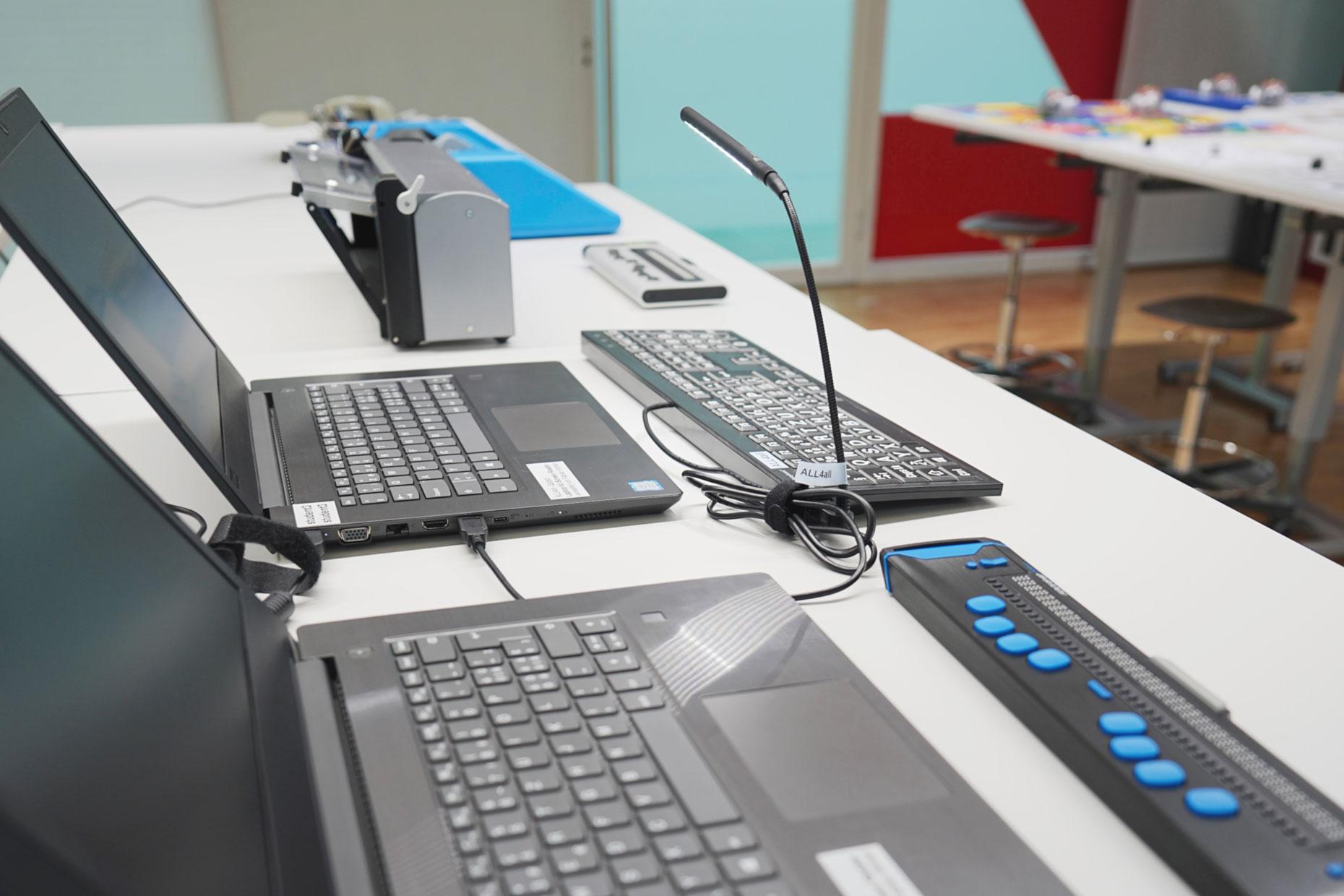 Foto eines Tisches mit aufgebauten Technologien des Bereichs Sehen. Auf dem Tisch ist folgendes aufgebaut; ein Laptop mit einer externen Tastatur mit grossen Tastenbeschriftungen und ein Laptop mit einer elektronischen Braillezeile.